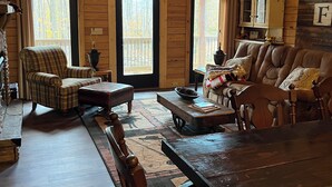 A spacious wooden cabin with a high ceiling, large windows, a cozy living area with a couch and chairs, a dining table, and a kitchen area with a cabinet.