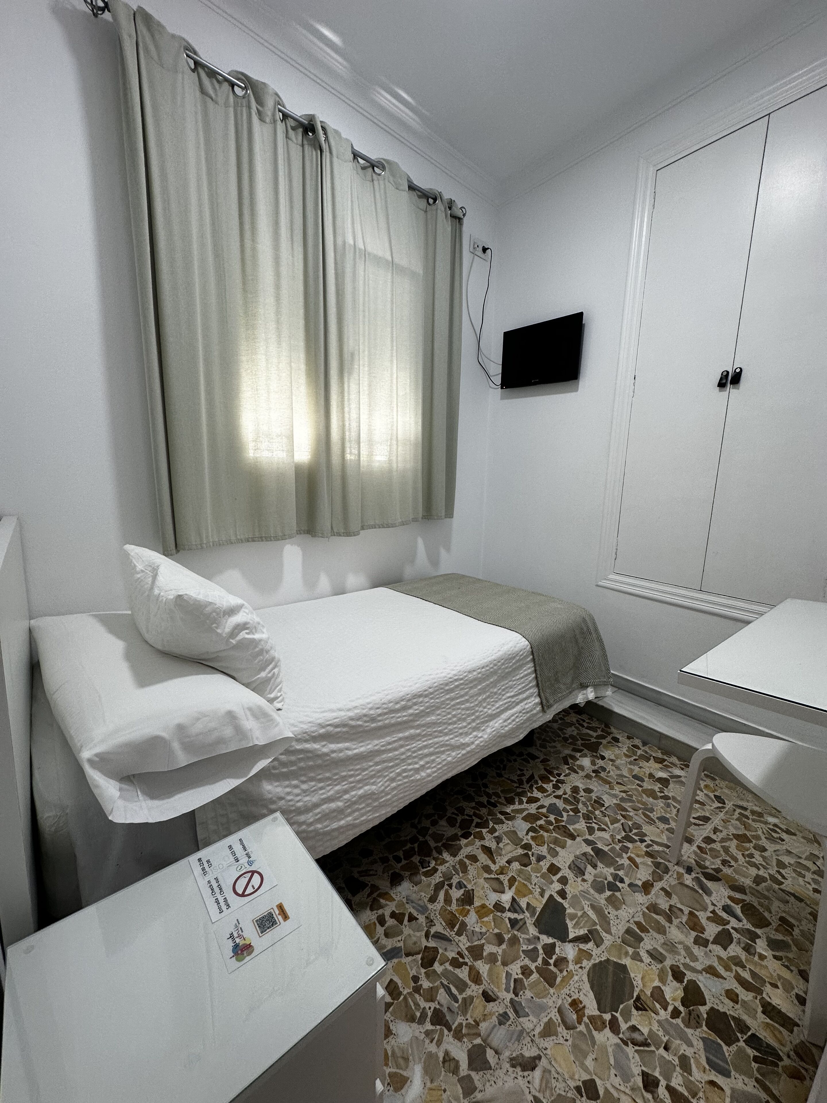 Una habitación pequeña y ordenada con una cama, una mesita, un televisor montado en la pared y un armario.