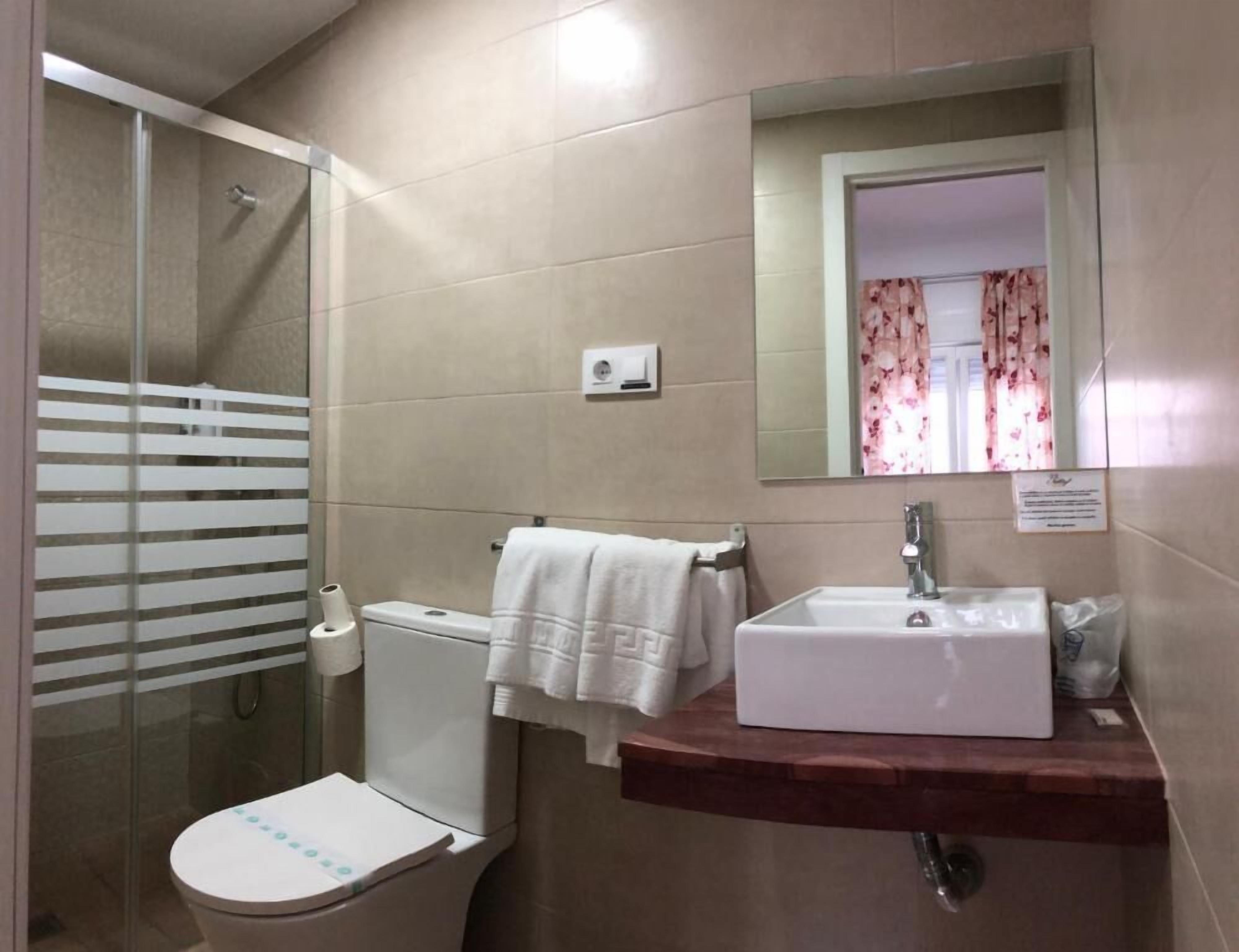 A bathroom with a glass shower enclosure, a white toilet, a sink with a wooden countertop, and a mirror with a floral curtain.