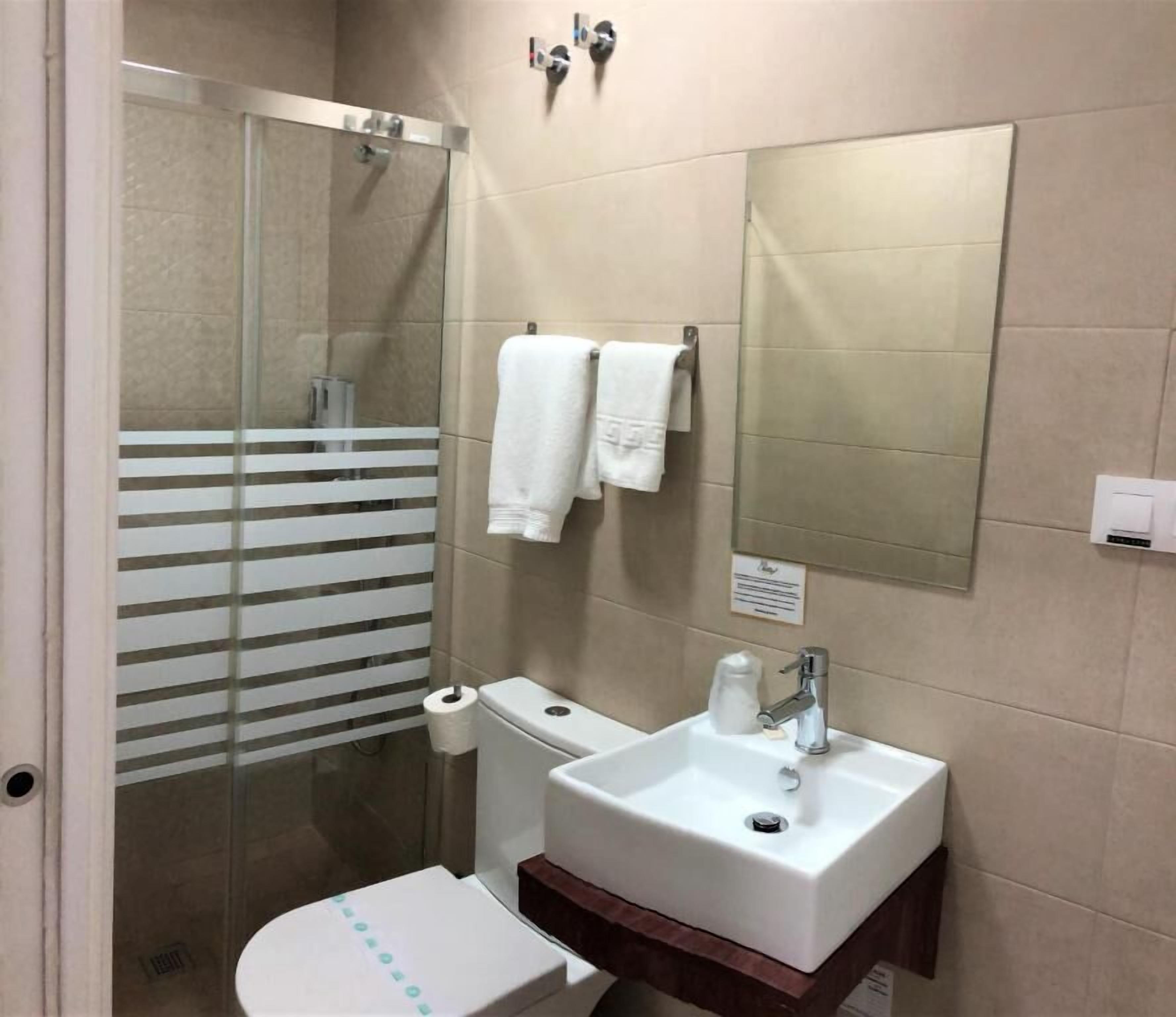 Un baño con cabina de ducha de vidrio, un lavamanos blanco con un espejo encima y dos toallas blancas colgadas en ganchos.