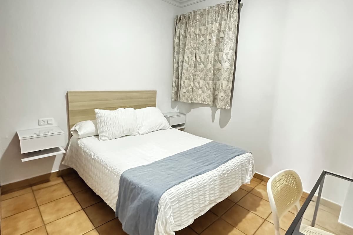 Une chambre à coucher avec un lit, une table de chevet, une chaise et une fenêtre avec des rideaux.