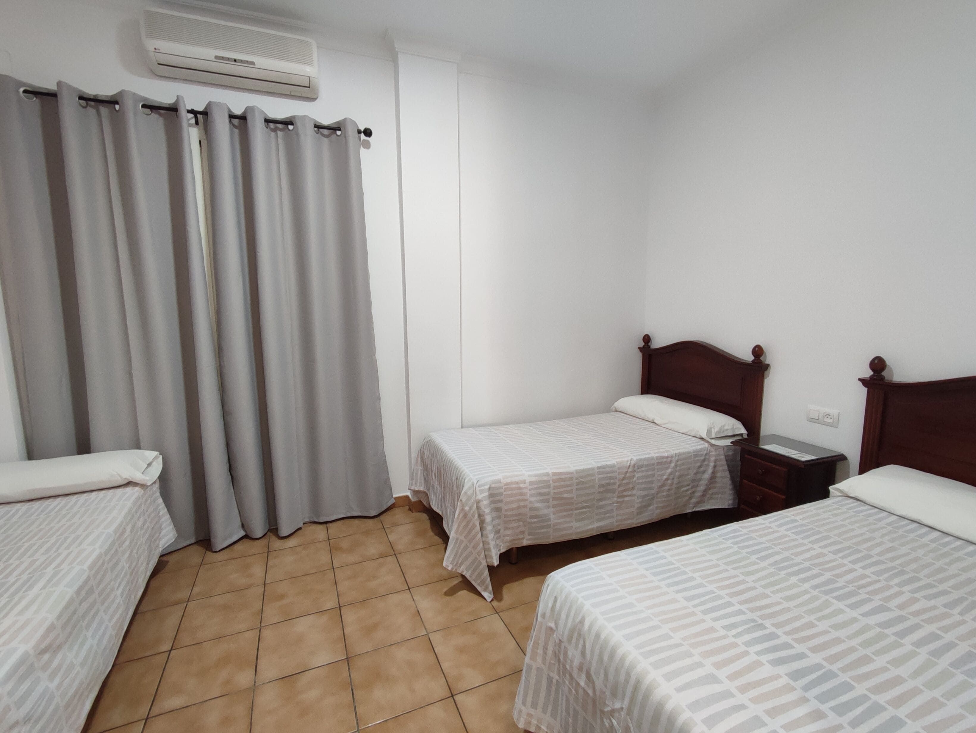 A bedroom with two single beds, a wooden nightstand, a white air conditioning unit, and a grey curtain.