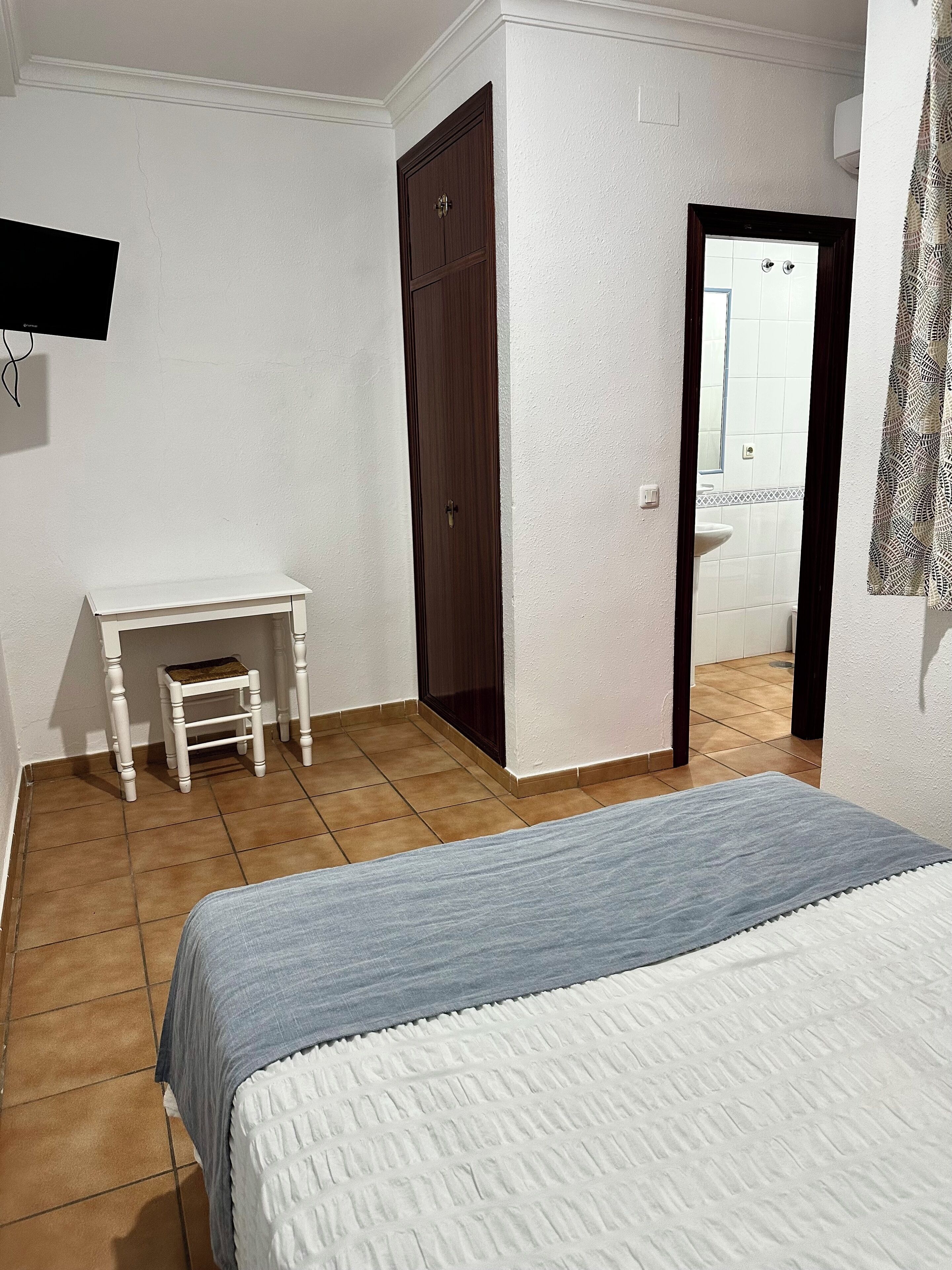 A bedroom with a bed, a small white desk, a wooden wardrobe, and a bathroom visible through an open door.
