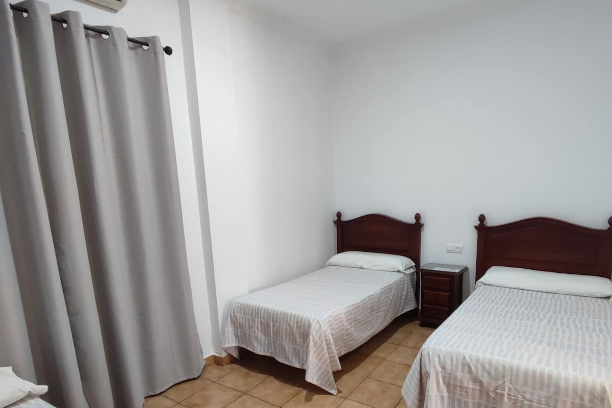 Une chambre avec deux lits simples, une petite table de chevet, un climatiseur fixé au mur et des rideaux gris.