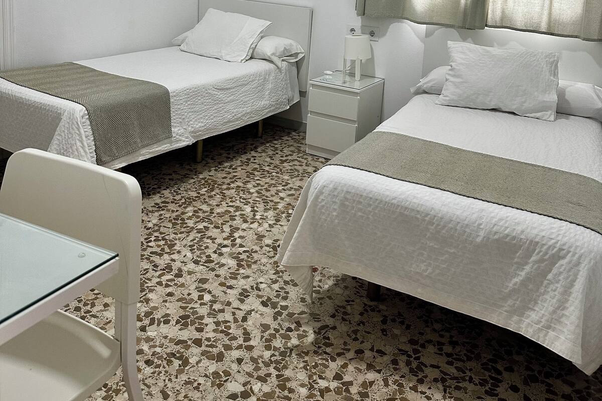 Une chambre d’hôtel avec deux lits, une petite table, une chaise, un appareil de climatisation et une fenêtre avec des rideaux.