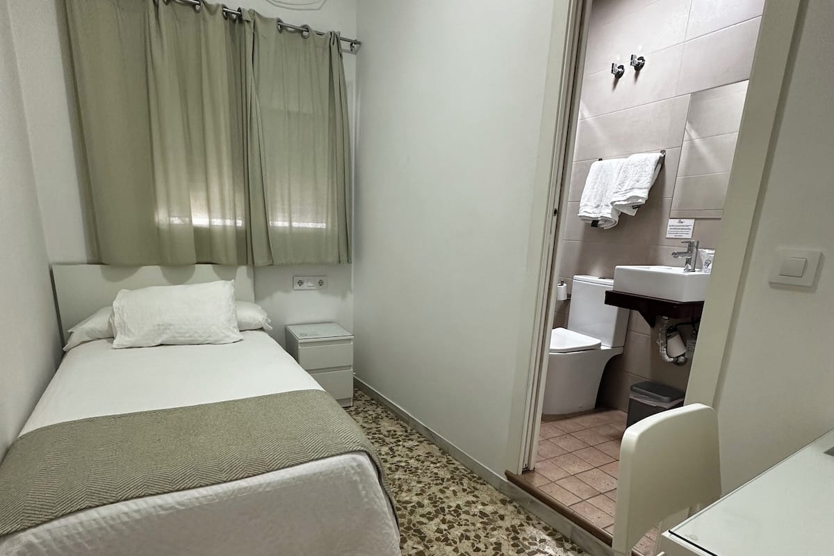 Une petite pièce propre, avec un lit, une chaise, un bureau et une salle de bain visible à travers une porte ouverte.