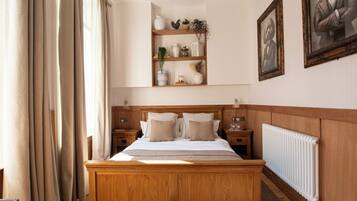 Quarto com cama de madeira, mesas de cabeceira, radiador e prateleira com objetos decorativos.