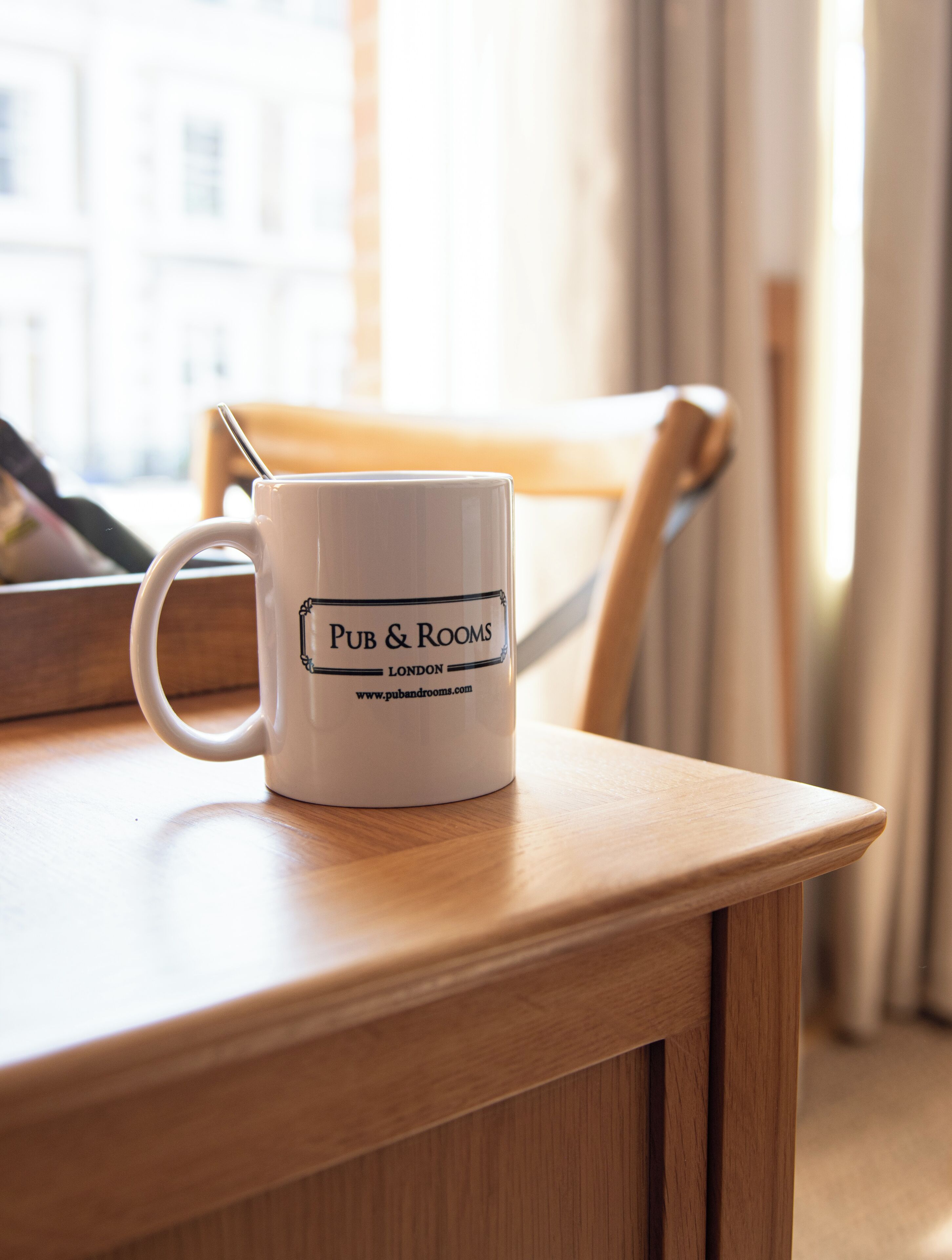 A white mug with "PUB & ROOMS LONDON" printed on it, placed on a wooden table with a chair in the background.