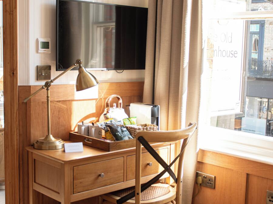 A wooden desk with a television, a lamp, and a chair. There is a window with curtains and a view of buildings outside.