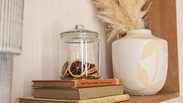 Uma prateleira de madeira com livros, um pote de vidro com fatias de laranja desidratada e um vaso com plumas de capim-do-campo.