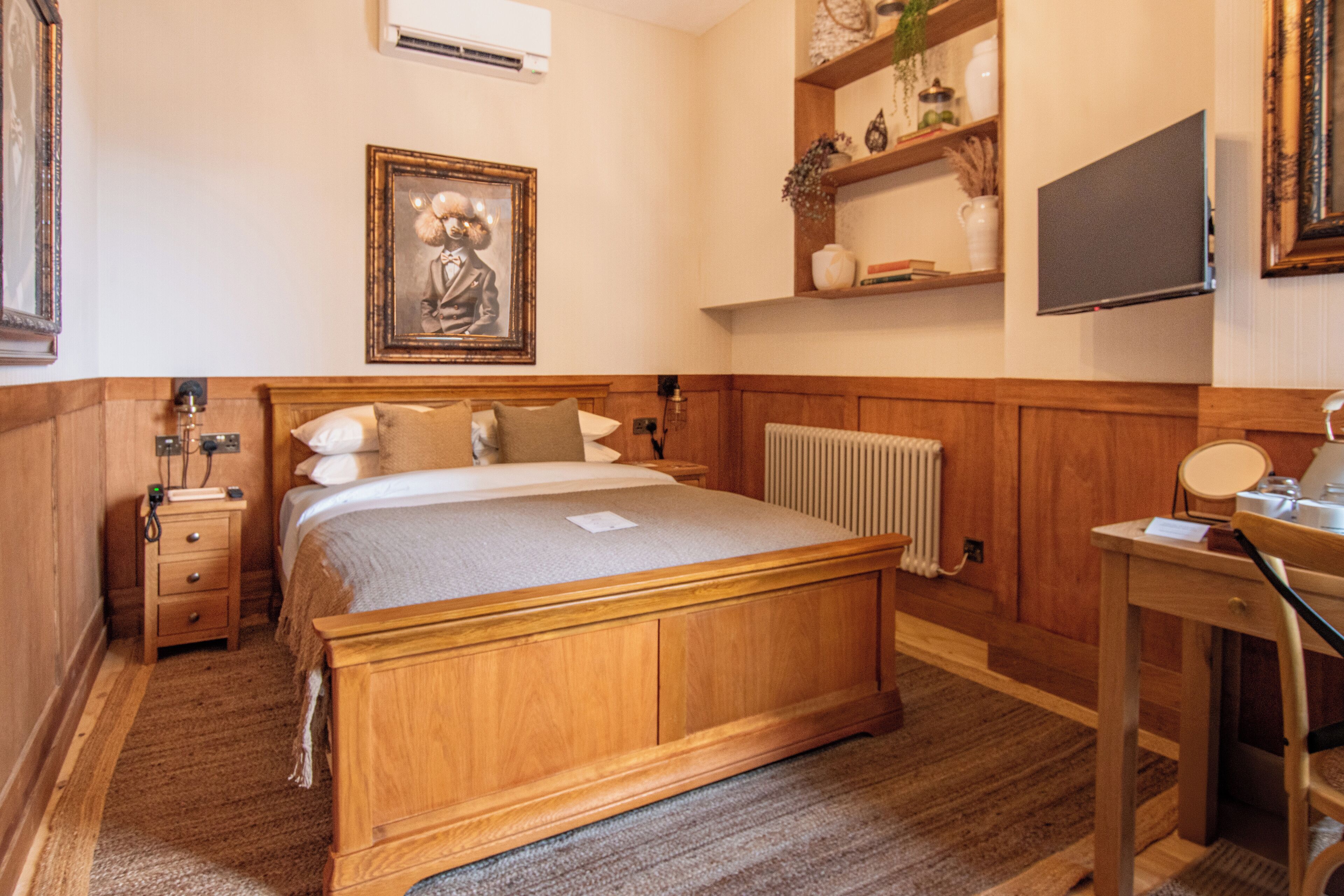 Un dormitorio con una cama de madera, una mesita de noche, un escritorio, una silla, un radiador, una estantería con objetos decorativos y un cuadro en la pared.