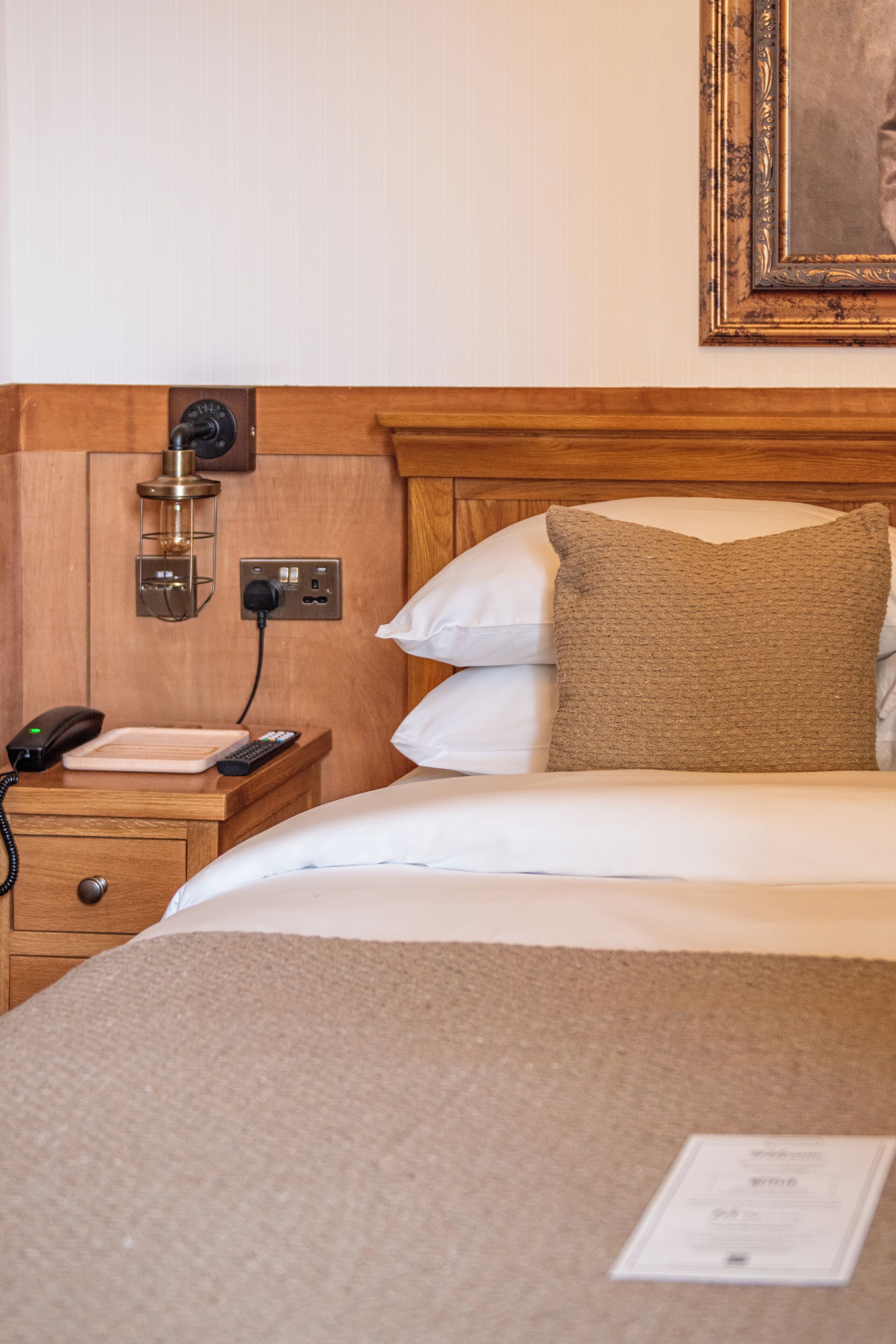 A hotel room with a wooden headboard, a bedside table with a phone and remote control, and a wall-mounted lamp.