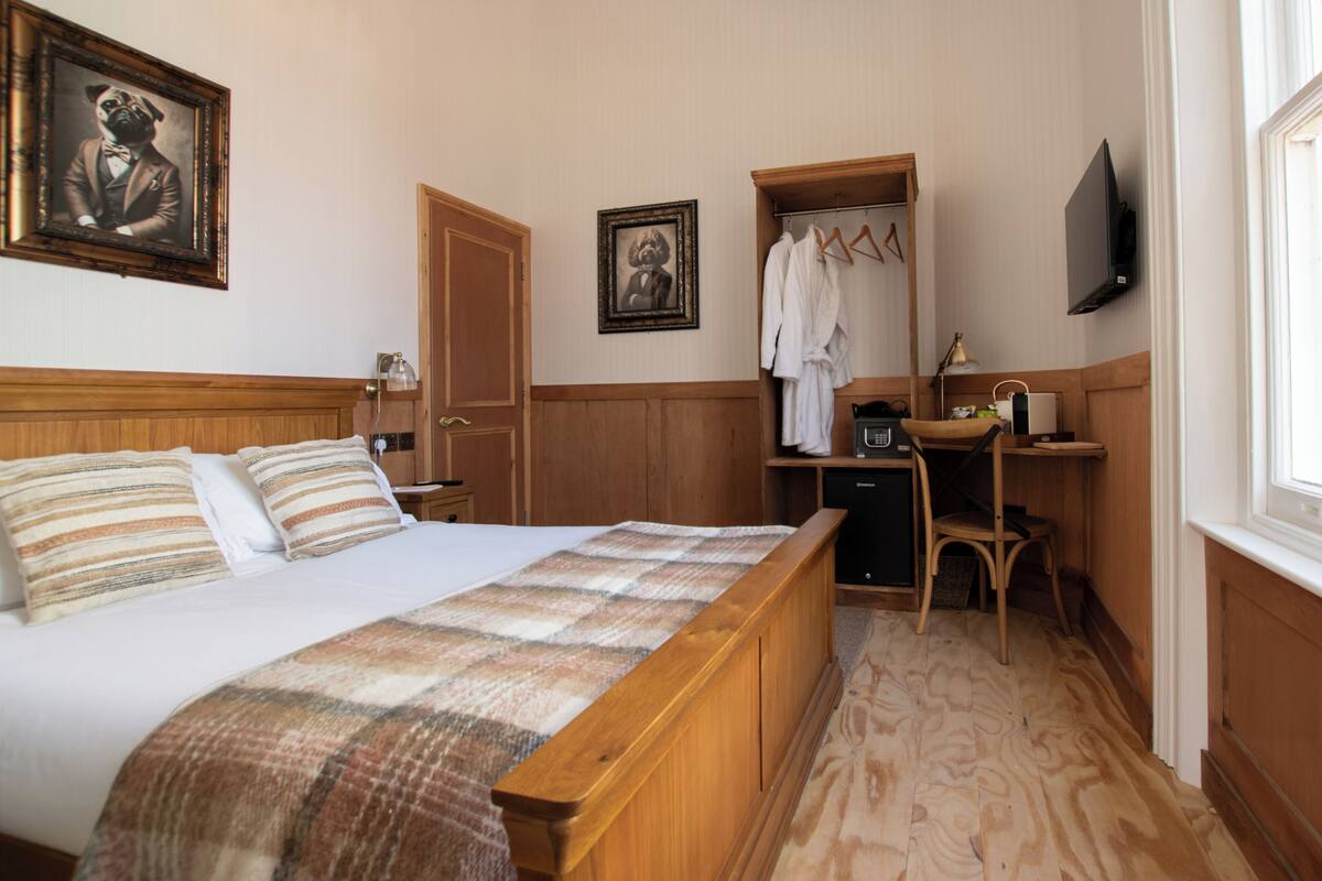 Une chambre d’hôtel avec un lit en bois, un bureau avec une chaise, un porte-manteau et un téléviseur fixé au mur.