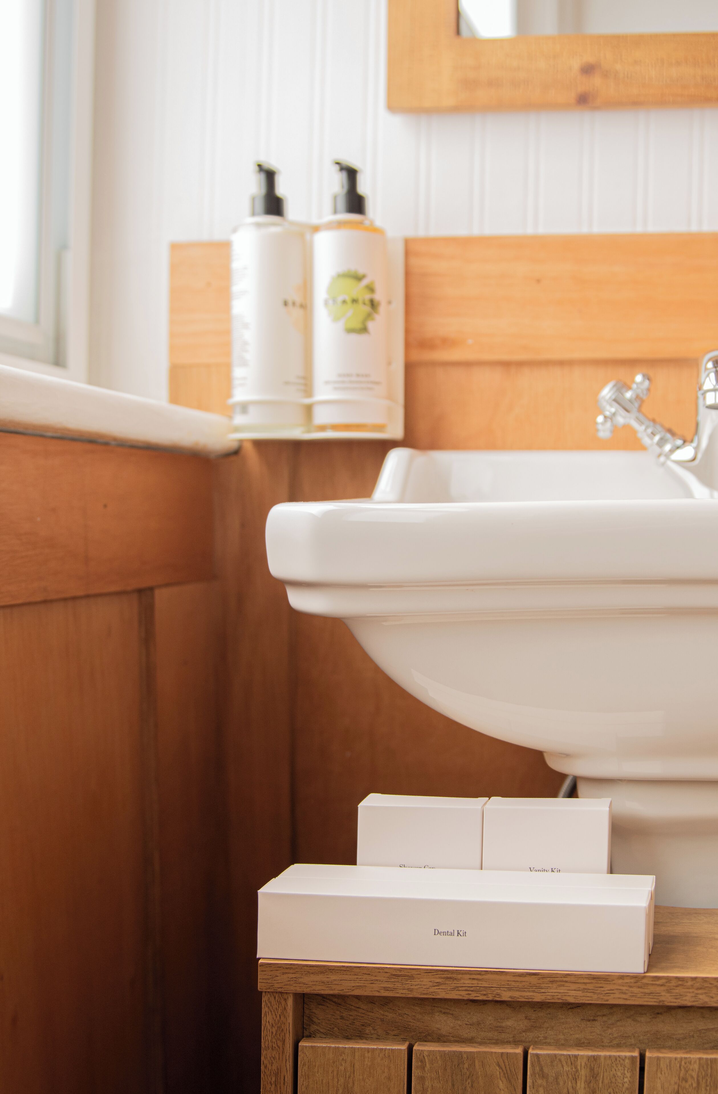 Une salle de bain avec un meuble-lavabo en bois, un lavabo blanc et deux flacons de produits de toilette sur l’étagère.