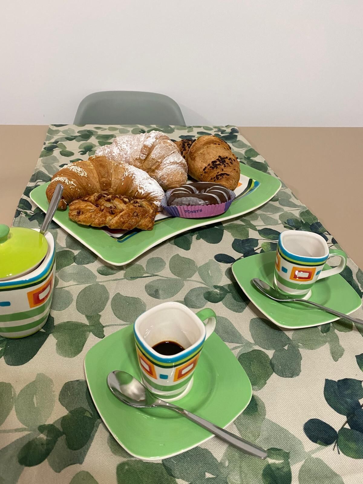 A table with pastries, coffee cups, and spoons on a patterned tablecloth.