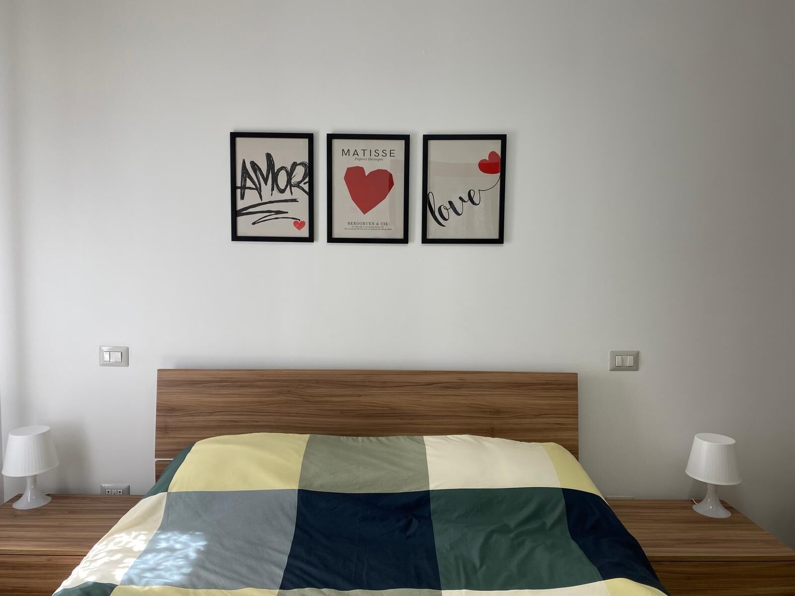 Une chambre à coucher avec une tête de lit en bois, une couvre-lit à carreaux et trois œuvres d’art encadrées accrochées au mur.