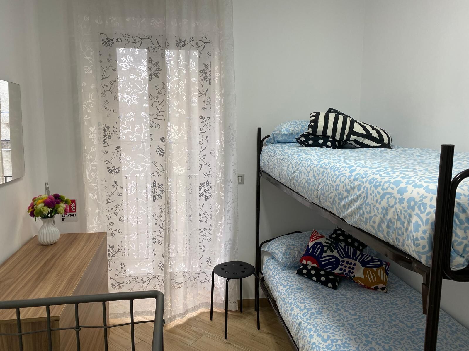 A bunk bed with patterned bedding, a small table with a vase of flowers, and a curtained window.
