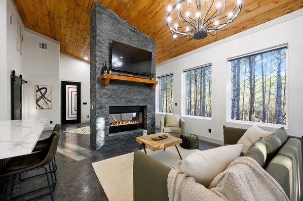Una sala de estar moderna con chimenea de piedra, un comedor y ventanas grandes con vista al bosque.