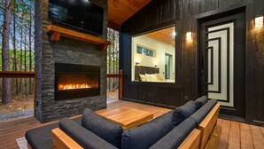 Un espacio al aire libre moderno con chimenea de piedra, mobiliario de madera y un amplio ventanal con vistas a un bosque.