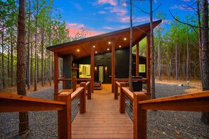 Guests can access the cabin's covered porch via a walkway with railings, surrounded by a forested environment.