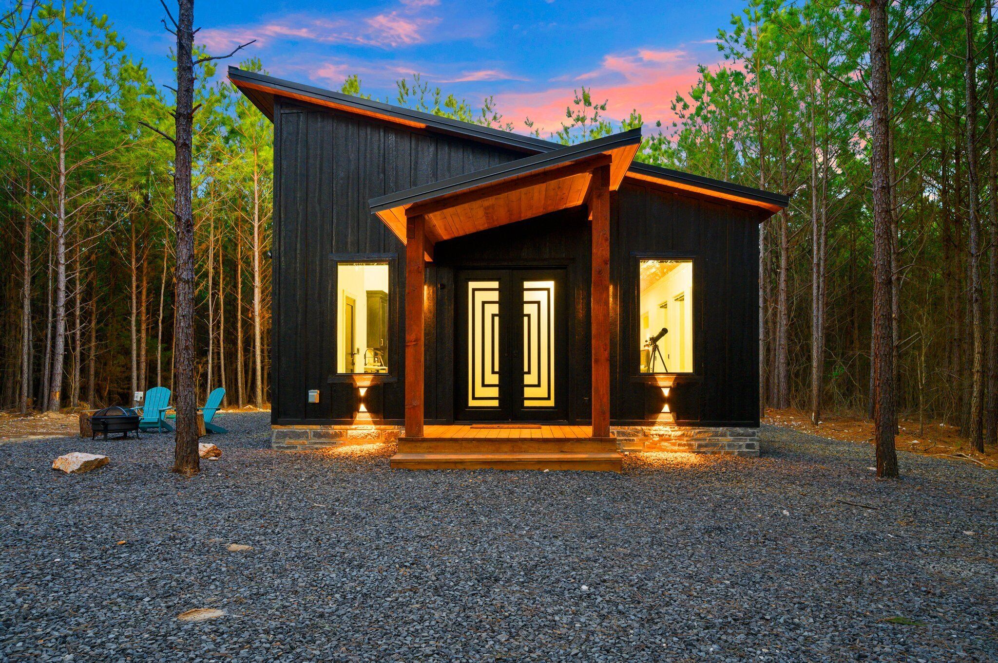 A modern cabin with a dark exterior, two large windows, and a wooden porch.