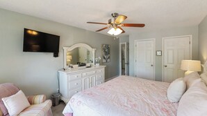 Un dormitorio con cama, cómoda, tocador, ventilador de techo y televisor montado en la pared.