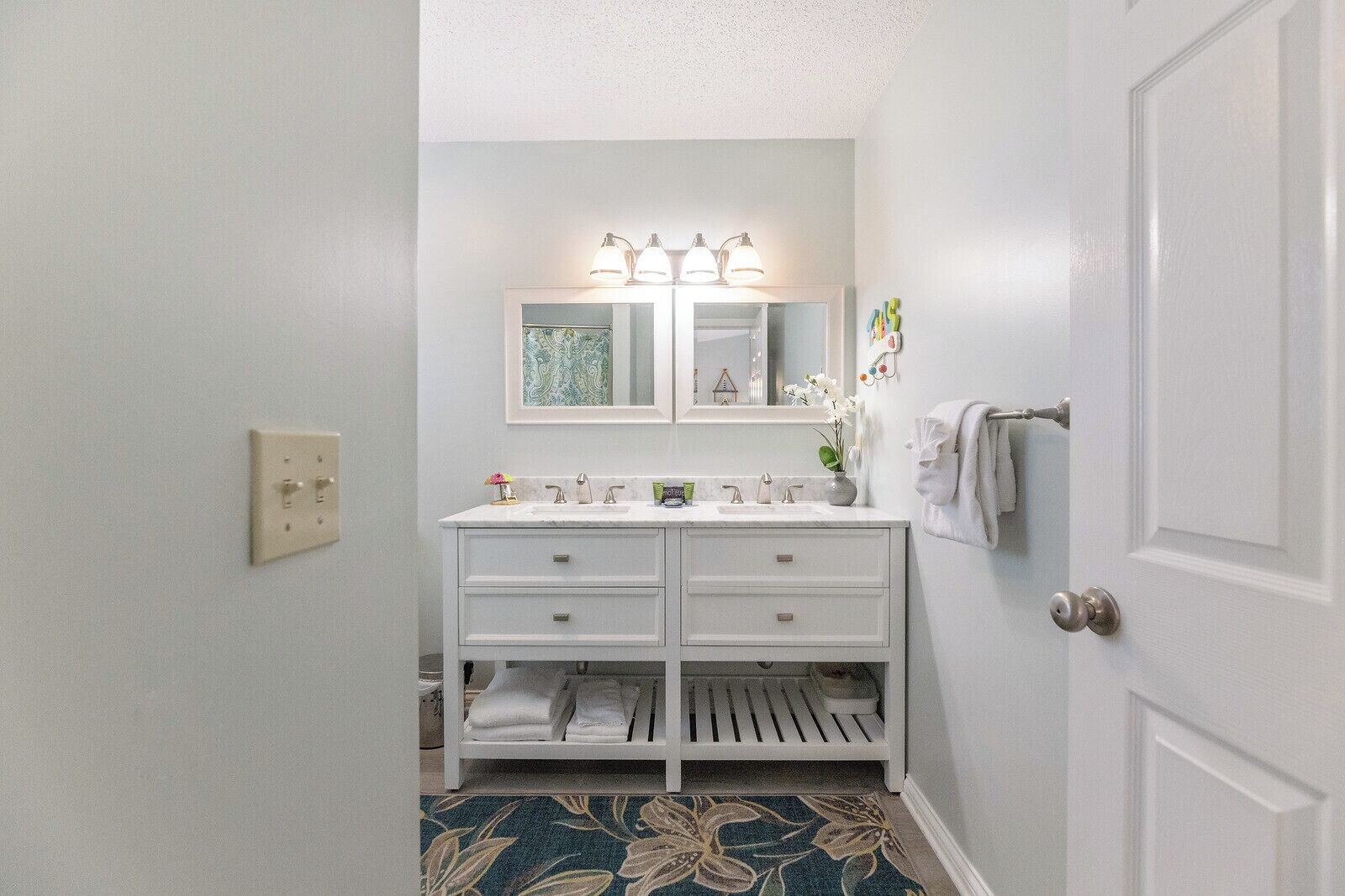 A bathroom with a white vanity, double sink, and a mirror with lights. There is a towel rack and a door to the right.