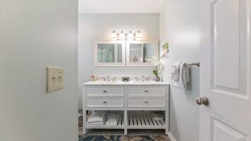 A bathroom with a white vanity, double sink, and a mirror with lights. There is a towel rack and a door to the right.