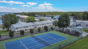 Una cancha de tenis con red, rodeada por una valla, cerca de un cuerpo de agua y edificios.