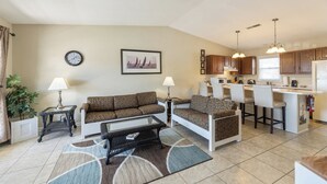 A living room with a sofa, a coffee table, and a dining area with a bar. There is a clock and a framed picture on the wall. - One Bedroom Harbor View Condo (Grand Rivers)