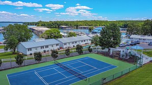 A tennis court surrounded by a fence, with a building and trees nearby. - One Bedroom Harbor View Condo (Grand Rivers)