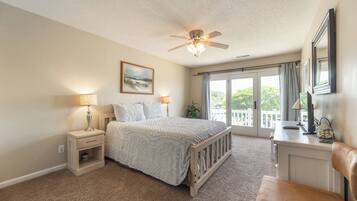 A bedroom with a bed, nightstands, a desk, a chair, a ceiling fan, and a large window with a view of greenery.