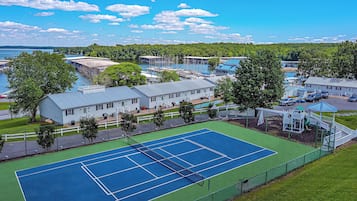 Un court de tennis entouré d’une clôture, avec un bâtiment et des arbres à proximité.