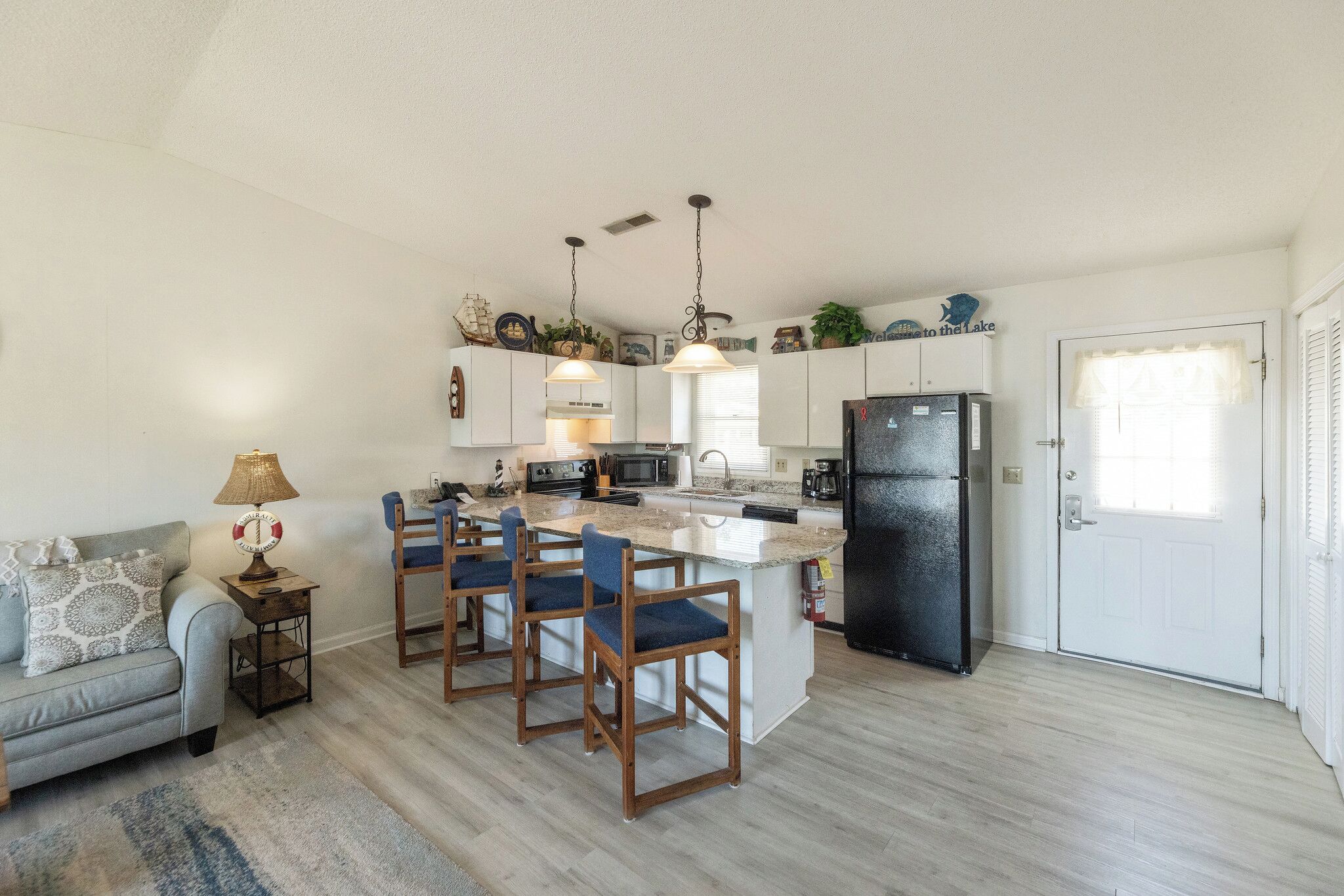 A compact kitchen with a central island, wooden chairs, a microwave, a refrigerator, and a dining area with a sofa and a lamp.