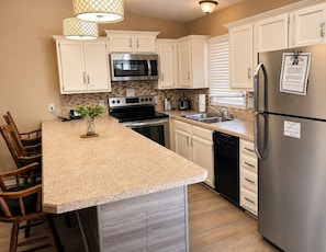 A kitchen with white cabinets, stainless steel appliances, a granite countertop, and a wooden dining set. - One Bedroom Harbor View Condo (Grand Rivers)