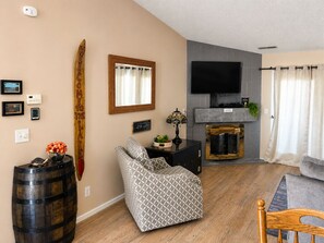 A living room with a TV, fireplace, patterned armchair, and wooden barrel. - One Bedroom Harbor View Condo (Grand Rivers)