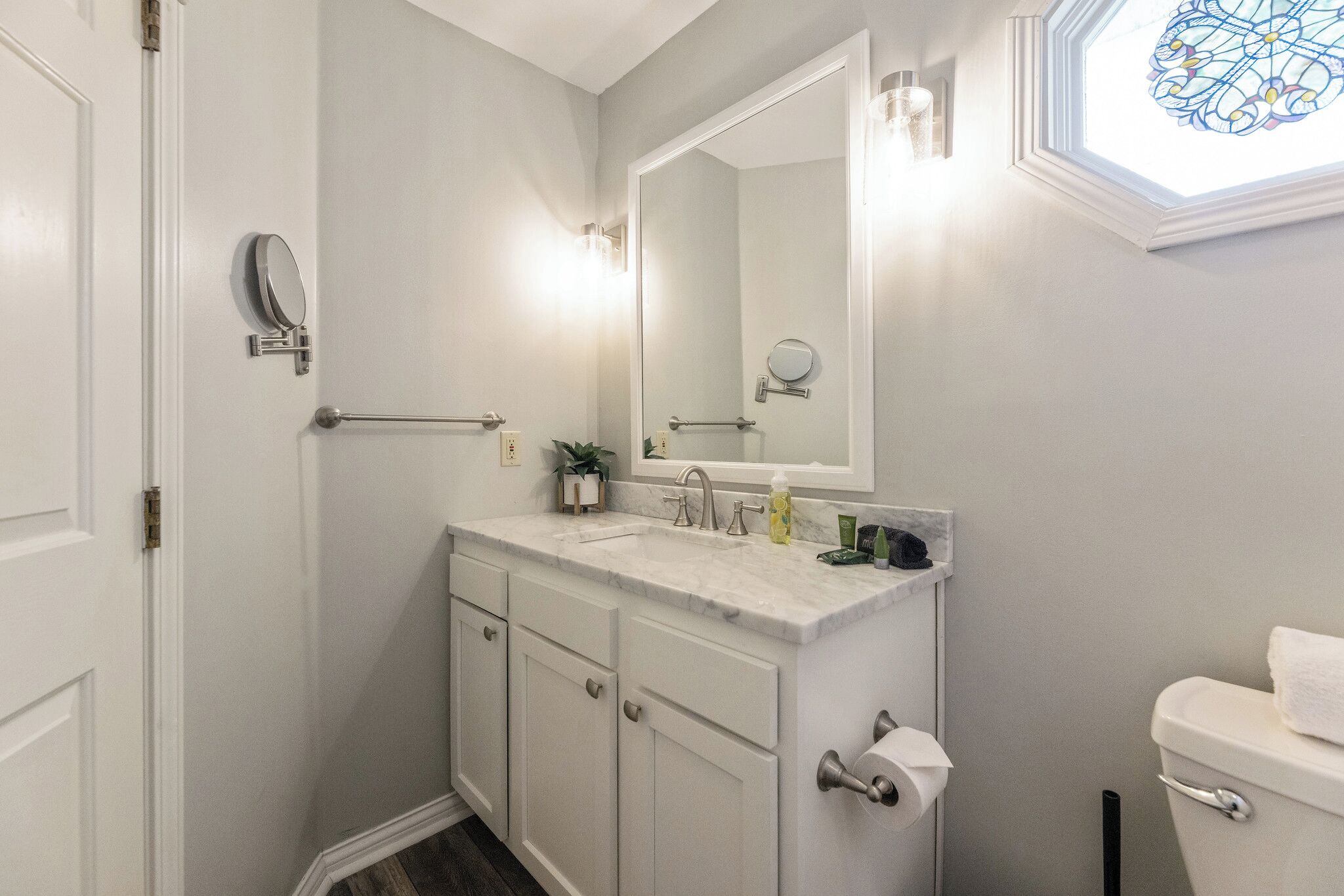 Une salle de bain avec un meuble-lavabo blanc, un grand miroir, des toilettes et une fenêtre ornée de vitraux.