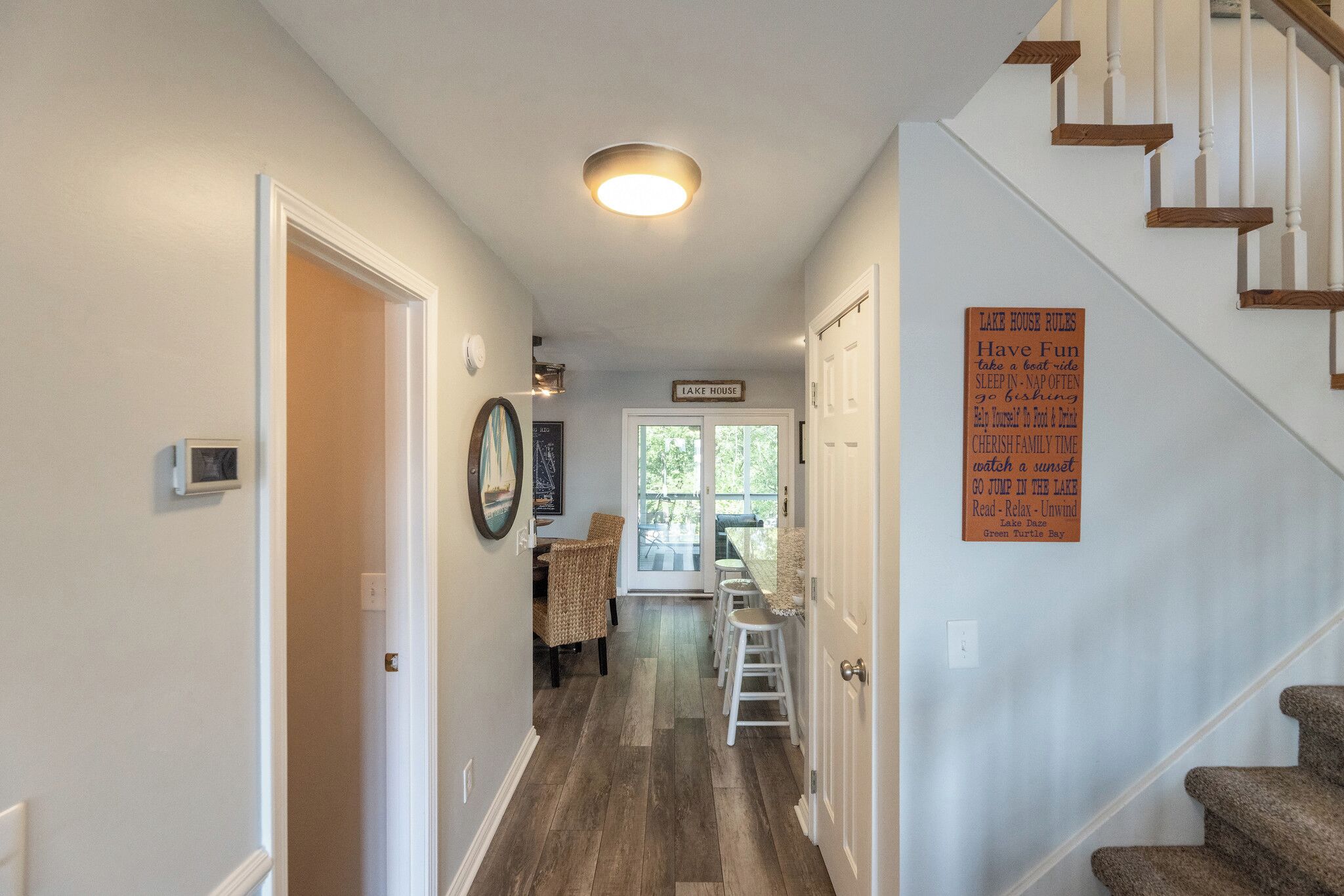 A hallway with a staircase, a dining area with wicker chairs, and a sign listing Lake House rules.