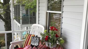 Une véranda avec une chaise blanche, une nappe à carreaux, un panneau et un arrangement floral.