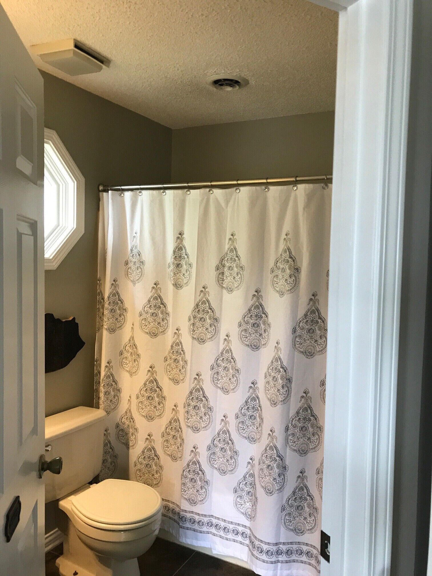 A bathroom with a toilet, a shower curtain with a paisley pattern, and a window above the toilet.