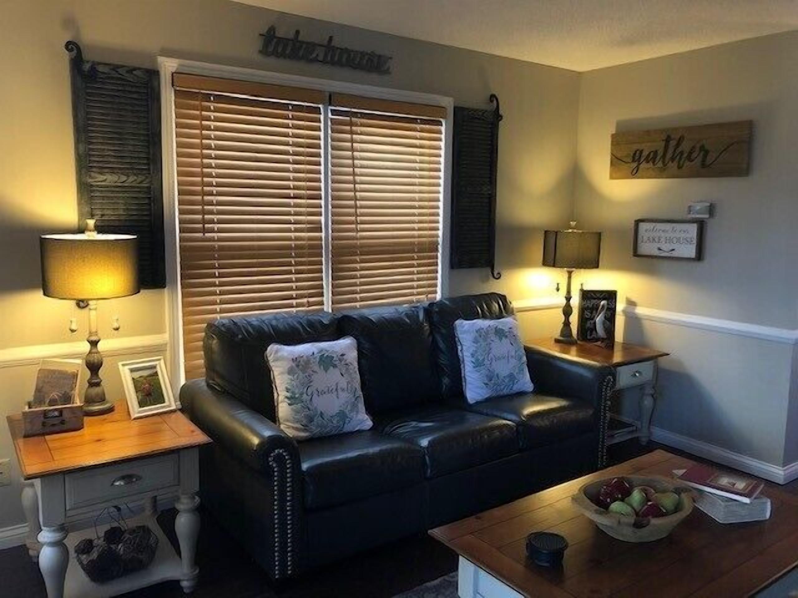 A living room with a black leather sofa, two lamps, a wooden coffee table, and a window with blinds.