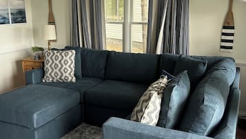 Un sofá modular con un reposapiés a juego, una mesita auxiliar con lámpara y vistas a través de una ventana con cortinas a rayas.