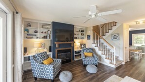 A living room with a fireplace, two patterned armchairs, a ceiling fan, and a staircase.