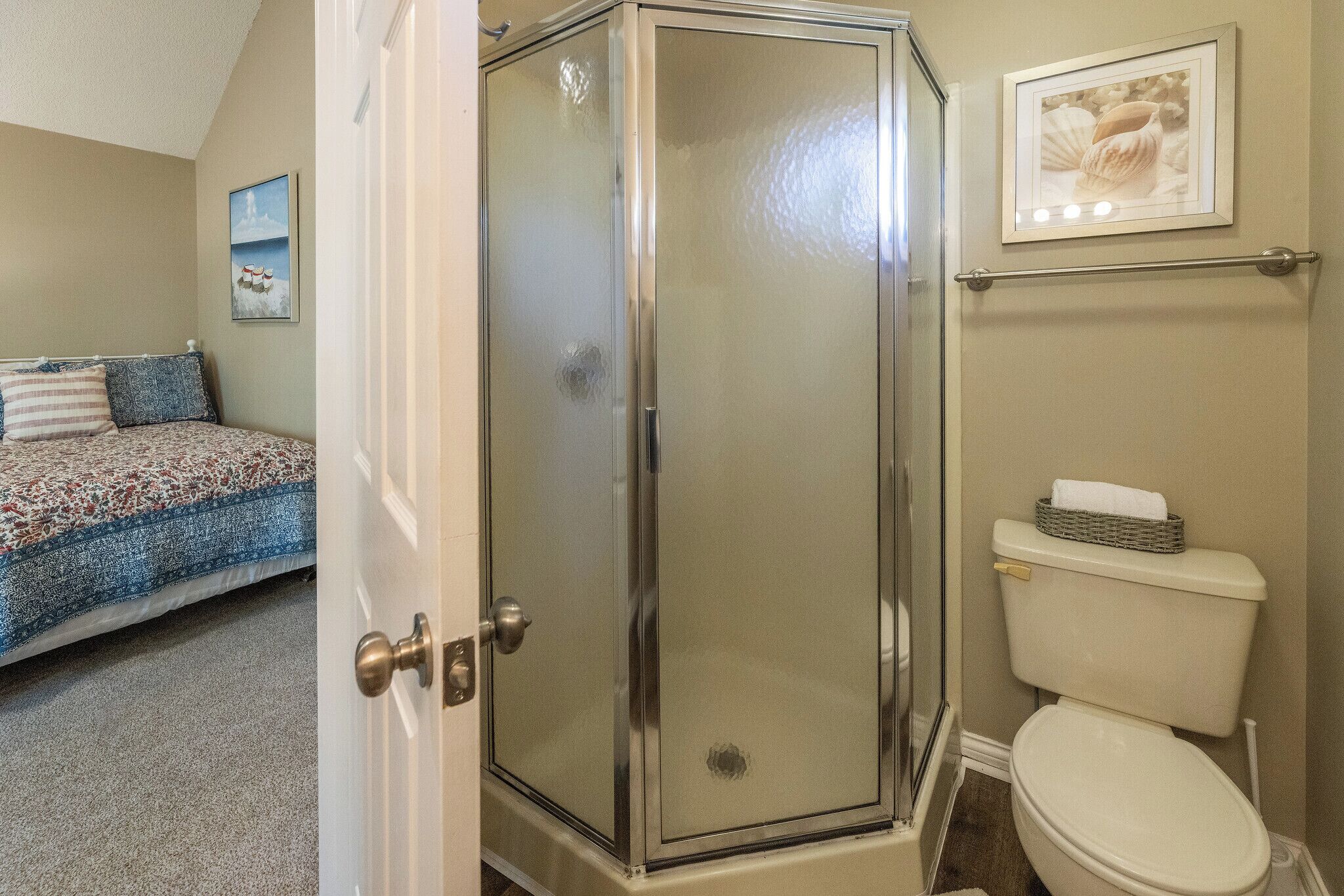 A bathroom with a glass-enclosed shower, a toilet, and a bed with a patterned bedspread.