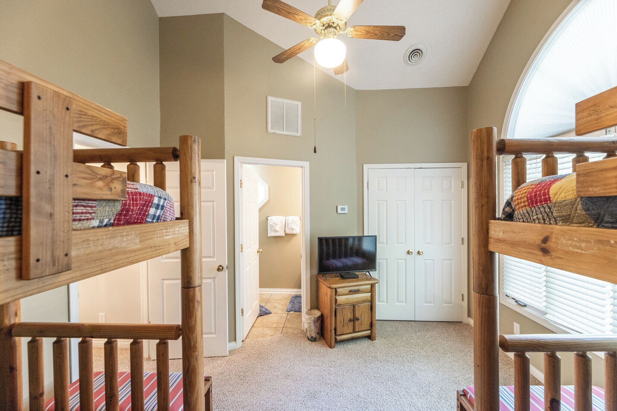 A room with bunk beds, a ceiling fan, a television on a wooden stand, and a door leading to another room.