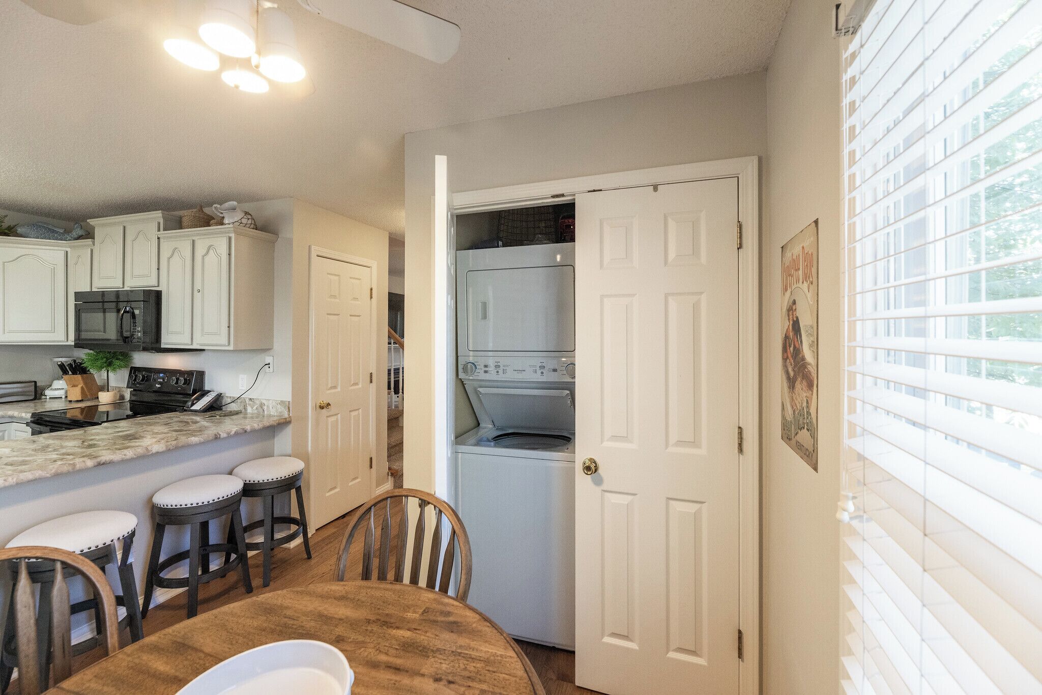 A kitchen with white cabinets, a built-in oven, and a washing machine. There is a dining table with stools and a window with blinds.