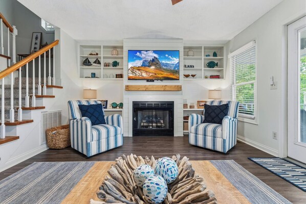 A living room with a fireplace, two striped armchairs, a television above the fireplace, and a wooden coffee table with decorative eggs.