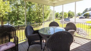 A covered patio with wicker furniture and a glass table, overlooking a parking area and trees.