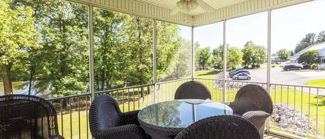 A covered patio with wicker furniture and a glass table, overlooking a parking area and trees.