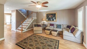 A spacious living room with a wooden staircase, a ceiling fan, a large framed wine bottle artwork, and a patterned rug.